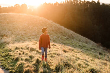 Kleiner Junge geht auf einem grasbewachsenen Hügel bei Sonnenuntergang - CAVF75939