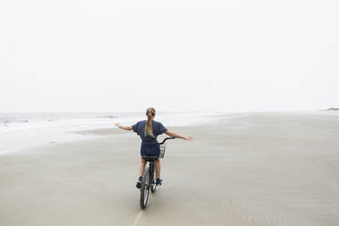 Ein junges Mädchen fährt mit dem Fahrrad an einem Sandstrand am Meer - MINF14053