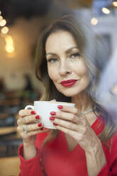 Portrait of mature woman with cup of coffee behind windowpane - PNEF02479