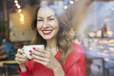Porträt einer lachenden reifen Frau mit einer Tasse Kaffee hinter einer Fensterscheibe - PNEF02480