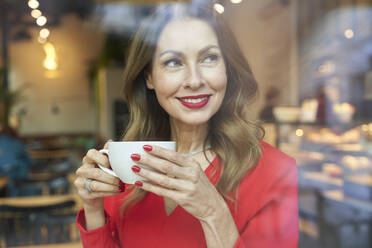 Porträt einer lächelnden reifen Frau mit einer Tasse Kaffee hinter einer Fensterscheibe - PNEF02482