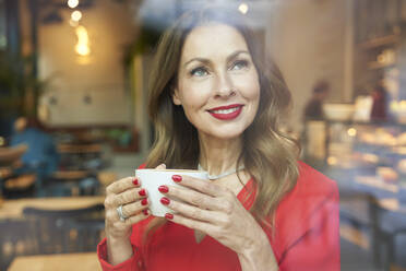 Porträt einer reifen Frau mit einer Tasse Kaffee hinter einer Fensterscheibe - PNEF02483