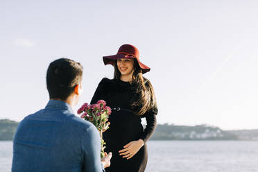 Junger Mann schenkt seiner schwangeren Freundin am Wasser Blumen - DCRF00050