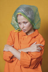 Porträt einer jungen Frau mit Plastiktüte auf dem Kopf, die eine orangefarbene Jacke vor einem gelben Hintergrund trägt - VPIF02115