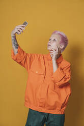 Portrait of young woman with short pink hair wearing orange jacket taking selfie with smartphone - VPIF02118