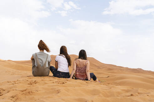 Rückansicht von drei jungen Frauen, die in einer Sanddüne in der Wüste Sahara sitzen, Merzouga, Marokko - AFVF05528
