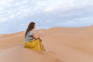 Junge Frau sitzt in einer Sanddüne in der Wüste Sahara und benutzt ein Handy, Merzouga, Marokko - AFVF05534