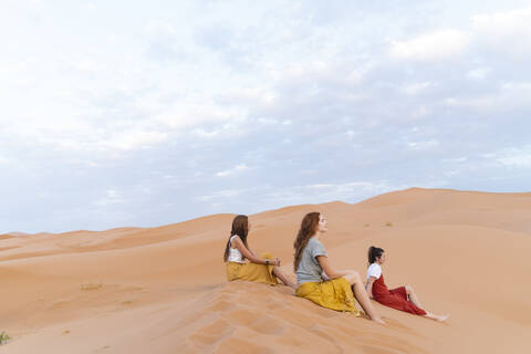 Drei junge Frauen sitzen auf einer Sanddüne in der Wüste Sahara, Merzouga, Marokko, lizenzfreies Stockfoto