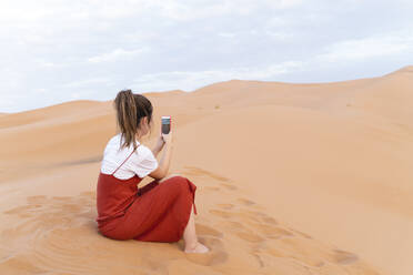 Junge Frau sitzt in einer Sanddüne in der Wüste Sahara und benutzt ein Handy, Merzouga, Marokko - AFVF05542