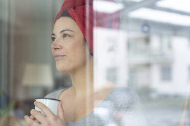 Porträt einer Frau mit in ein Handtuch gewickeltem Kopf, die hinter einer Fensterscheibe einen Kaffee trinkt - MOEF02852