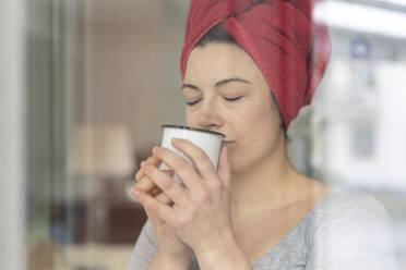Porträt einer Frau mit in ein Handtuch gewickeltem Kopf, die hinter einer Fensterscheibe einen Kaffee trinkt - MOEF02853