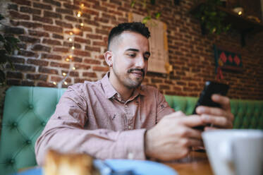 Porträt eines lächelnden Mannes in einem Café, der auf sein Mobiltelefon schaut - KIJF02942