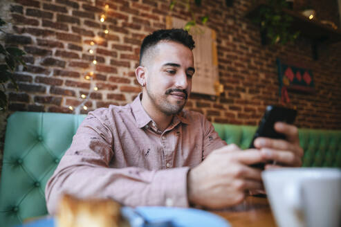 Porträt eines lächelnden Mannes in einem Café, der auf sein Mobiltelefon schaut - KIJF02942