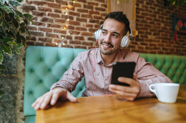 Lächelnder Mann in einem Cafe, der mit Kopfhörern Musik hört - KIJF02946