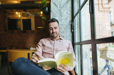 Mann liest Buch in einem Café am Fenster - KIJF02950