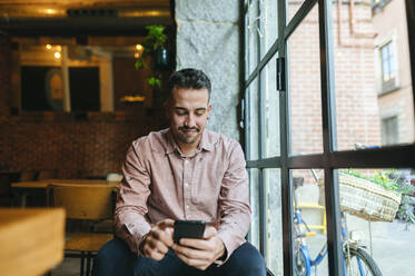 Mann am Fenster eines Cafés schaut auf sein Handy - KIJF02952