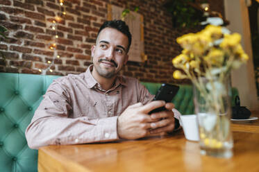 Lächelnder Mann in einem Café, der ein Mobiltelefon hält - KIJF02953