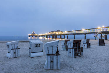 Germany, Mecklenburg-Western Pomerania, Heringsdorf, Hooded beach chairs on sandy coastal beach at dusk with illuminated pier in background - WDF05857