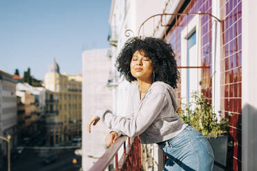 Porträt einer jungen Frau mit lockigem Haar, die auf einem Balkon steht und die Sonne genießt - DCRF00078