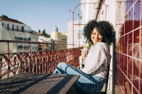Glückliche junge Frau mit lockigem Haar sitzt auf dem Balkon und genießt die Sonne - DCRF00079