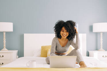 Glückliche junge Frau mit lockigem Haar, die einen Laptop auf dem Bett zu Hause benutzt - DCRF00088