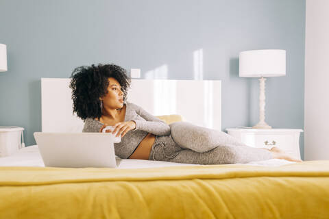 Junge Frau mit Kaffeetasse und Laptop zu Hause auf dem Bett liegend, lizenzfreies Stockfoto