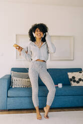 Happy young woman with curly hair listening to music and dancing in living room at home - DCRF00096