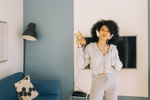 Happy young woman with curly hair listening to music and dancing in living room at home stock photo