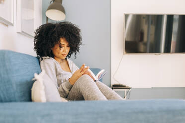 Young woman with curly hair reading a book on sofa at home - DCRF00098