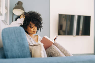 Junge Frau mit lockigem Haar liest ein Buch auf dem Sofa zu Hause - DCRF00099