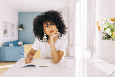 Portrait of young woman with curly hair sitting at a table with a notebook at home - DCRF00101