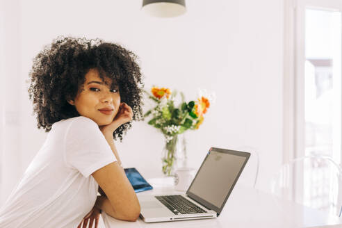 Porträt einer selbstbewussten jungen Frau mit lockigem Haar, die einen Laptop auf einem Tisch zu Hause benutzt - DCRF00103