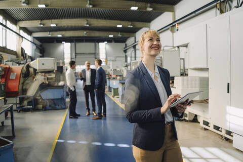 Porträt einer lächelnden Geschäftsfrau mit Tablet in einer Fabrik mit Kollegen im Hintergrund, lizenzfreies Stockfoto