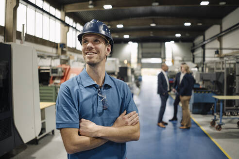 Porträt eines selbstbewussten Arbeiters in einer Fabrik mit Schutzhelm und Geschäftsleuten im Hintergrund - KNSF07706