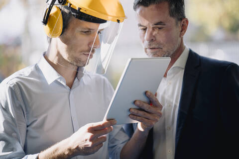 Geschäftsmann und Mann mit Schutzhelm schauen auf ein Tablet, lizenzfreies Stockfoto