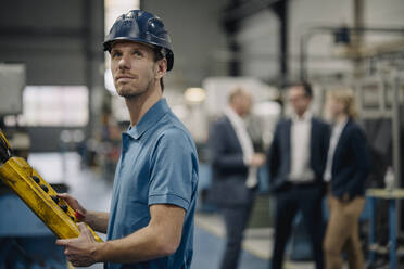 Arbeiter in einer Fabrik, der eine Maschine bedient, mit Geschäftsleuten im Hintergrund - KNSF07737