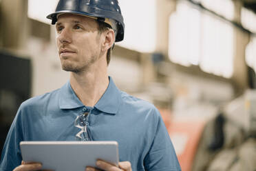 Porträt eines Arbeiters in einer Fabrik, der einen Schutzhelm trägt und ein Tablet hält - KNSF07739