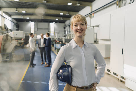 Portrait of a smiling woman with hard hat in a factory with colleagues in backgound stock photo