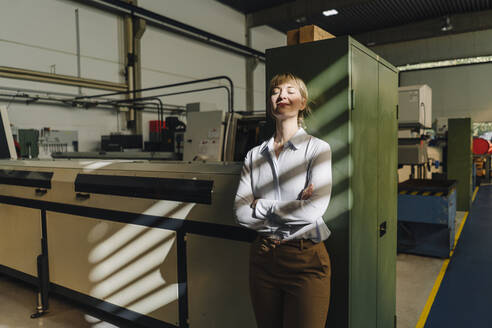 Businesswoman with closed eyes leaning against a cabinet in a factory - KNSF07785