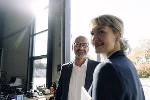 Porträt eines lächelnden Geschäftsmanns und einer Geschäftsfrau am Fenster in einer Fabrik - KNSF07804