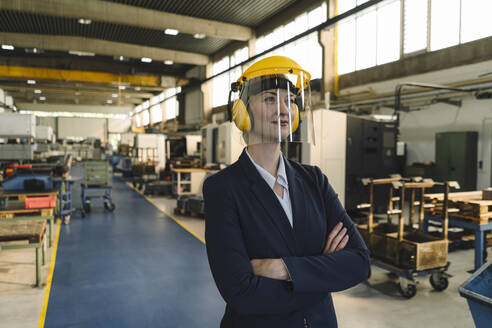 Portrait of a confident businesswoman wearing safety helmet and earmuffs in a factory - KNSF07809