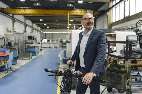 Lächelnder Geschäftsmann mit Fahrrad in einer Fabrik, lizenzfreies Stockfoto