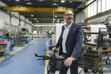 Lächelnder Geschäftsmann mit Fahrrad in einer Fabrik - KNSF07827