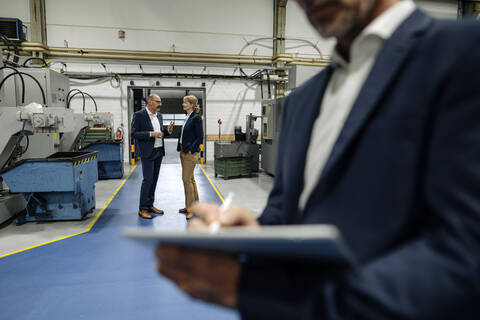 Geschäftsmann, der ein Tablet in einer Fabrik benutzt, mit Kollegen, die sich im Hintergrund unterhalten, lizenzfreies Stockfoto