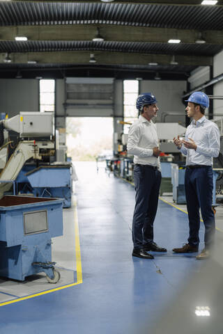 Zwei Männer mit Schutzhelmen unterhalten sich in einer Fabrik, lizenzfreies Stockfoto