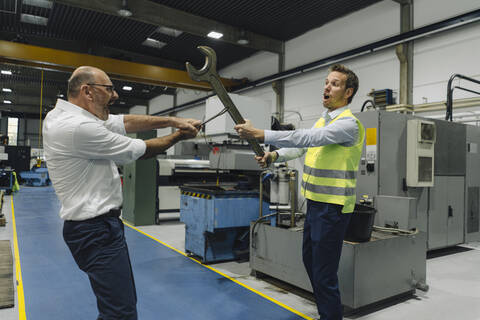 Geschäftsmann und Mann in reflektierender Weste liefern sich in einer Fabrik einen Kampf mit großen Schraubenschlüsseln, lizenzfreies Stockfoto