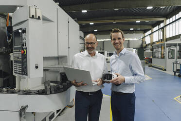 Porträt von zwei lächelnden Männern in einer Fabrik mit Laptop und VR-Brille - KNSF07870