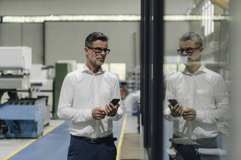 Älterer Geschäftsmann mit Mobiltelefon in einer Fabrik, die sich in einer Glasscheibe spiegelt - KNSF07911