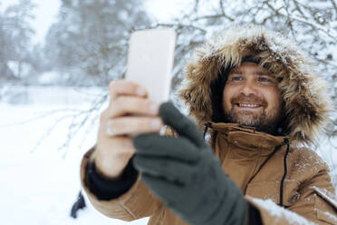 Portrait of smiling man taking selfie with with cell phone in winter - KNTF04426