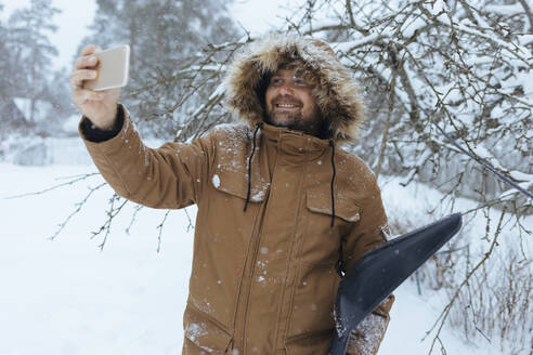 Porträt eines lächelnden Mannes mit Schneeschaufel, der ein Selfie mit seinem Mobiltelefon macht - KNTF04427